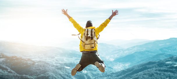Osoba w żółtej kurtce sportowej z plecakiem podskakuje na tle górskiego pejzażu.
