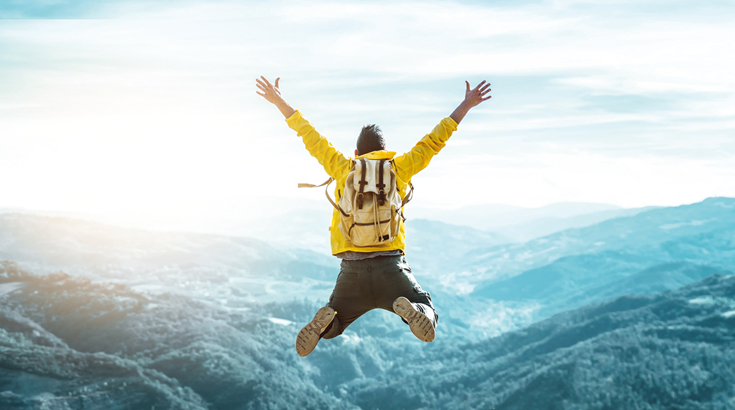 Osoba w żółtej kurtce sportowej z plecakiem podskakuje na tle górskiego pejzażu.