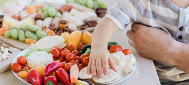 Ręka małego dziecka sięga po jedzenie na talerzu pełnym warzyw i serów, w tle stół zastawiony owocami