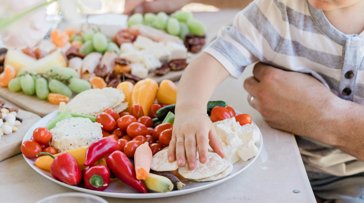 Ręka małego dziecka sięga po jedzenie na talerzu pełnym warzyw i serów, w tle stół zastawiony owocami