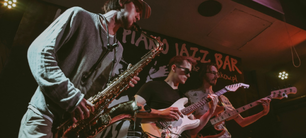 Trzech muzyków z zespołu Szymon gra na scenie klubu jazzowego, uwaga skupia się na saksofoniście (po lewej), obok dwóch gitarzystów, wszyscy grają.