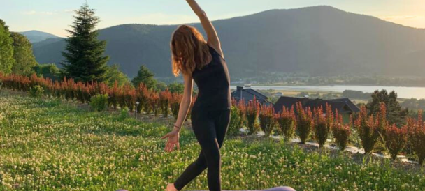 kobieta stoi na macie ułożonej w trawie, w pozycji z jogi, z jedną ręką wyprostowaną i unisioną nad głową. W tle górski krajobraz.