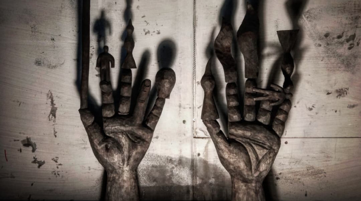 Grafika promująca wystawę Dowód Rzeczowy, dwie wyrzeźbione dłonie rzucające cień na ścianę, na palcach rzeźby różnych kształtów - zdjęcie ma niepokojący nastrój