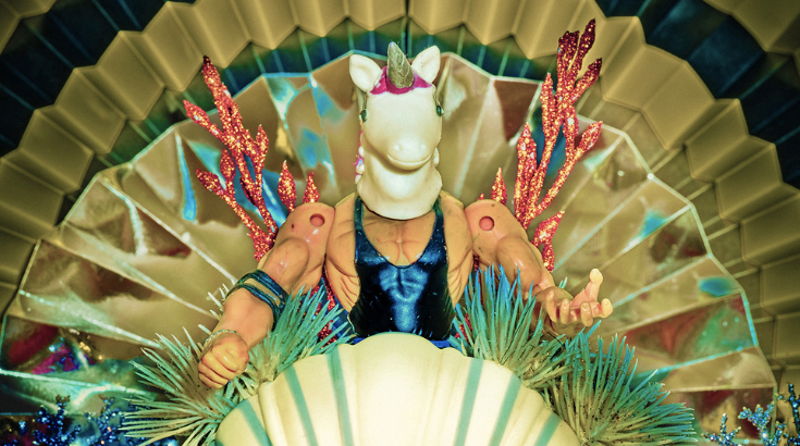 Grafika promująca wystawę "Odbicia" - z przodu, za muszlą przypominającą didżejkę, stoi lalka-ken z głową jednorożca, otoczony symetrycznymi, plastikowymi roślinami, z tyłu złote tło przypominające rozłożony wachlarz