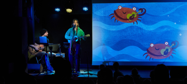 Zdjęcie ze spektaklu "O krabie Heniu". Dwie osoby z gitarami, jedna śpiewająca przy mikrofonie. Obok projektor z animacją z dwoma krabami grającymi na instrumentach muzycznych.