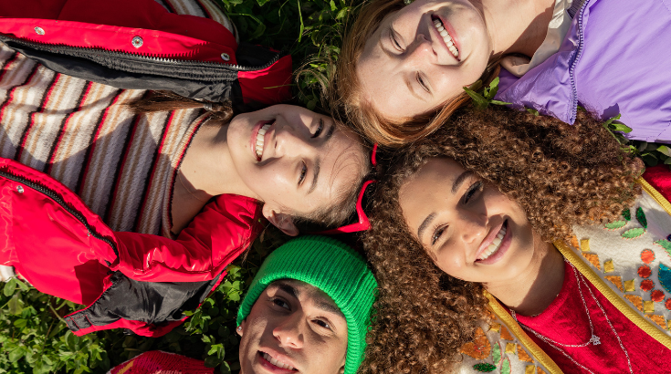 Czwórka młodych osób leżąca w trawie stykając się głowami, z uśmiechami na twarzach