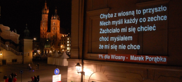 Na pierwszym planie widać wyświetlony na ścianie budynku tekst wiersza. W tle po lewej stronie Kościół Mariacki.