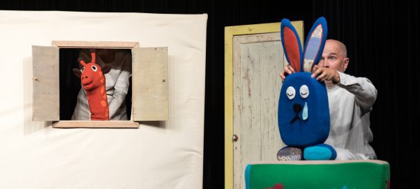 scena teatralna aktor porusza postacią niebieskiego królika obok pluszowa czerwona żyrafa