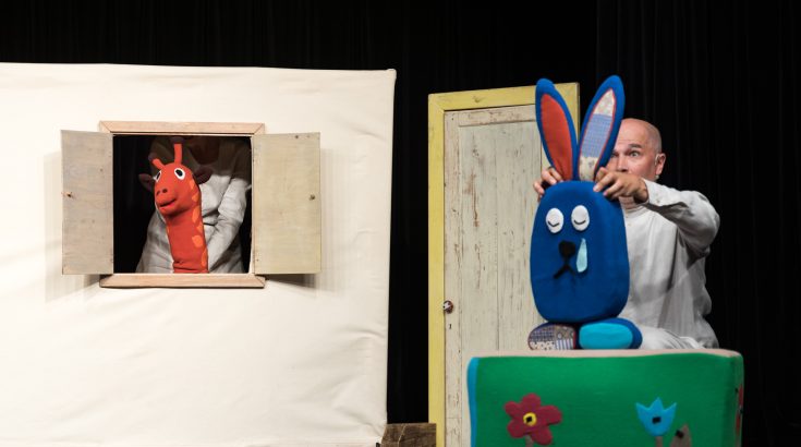 scena teatralna aktor porusza postacią niebieskiego królika obok pluszowa czerwona żyrafa