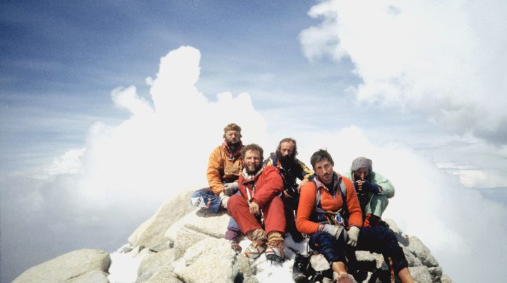 Pięcioro mężczyzn, himalaistów, siedzi na szczycie góry. W tle chmury.