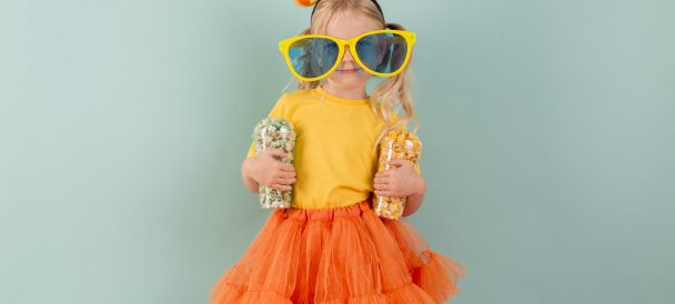 Na zielonym tle mała dziewczynka ubrana na żółto - pomarańczowo. W rękach trzyma kubki z cukierkami, na twarzy ma wielkie, żółte okulary a na głowie dużą kokardę w kropki.