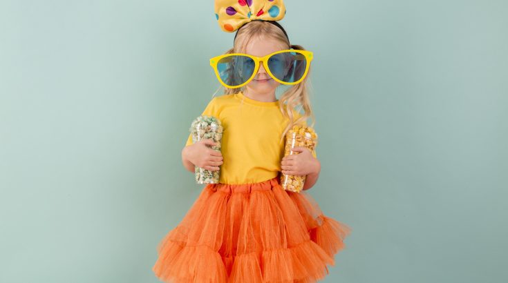 Na zielonym tle mała dziewczynka ubrana na żółto - pomarańczowo. W rękach trzyma kubki z cukierkami, na twarzy ma wielkie, żółte okulary a na głowie dużą kokardę w kropki.