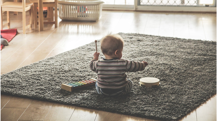 Odwrócone tyłem małe dziecko siedzi na dywanie i gra na cymbałkach i bębenku.