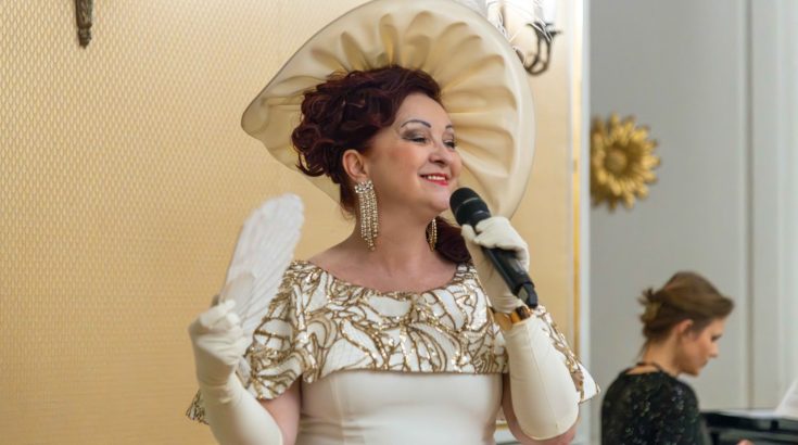 Śpiewaczka operowa w biało-złotym stroju i kapeluszu z białym wachlarzem.