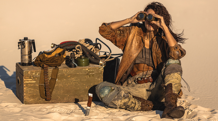 Dziewczyna siedząca na piasku z lornetką, obok skrzynia drewniana.