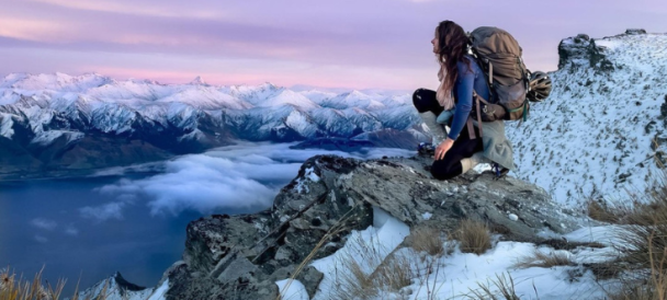 Na pierwszym planie na skale siedzi kobieta z plecakiem turystycznym wpatrująca się w krajobraz górski.