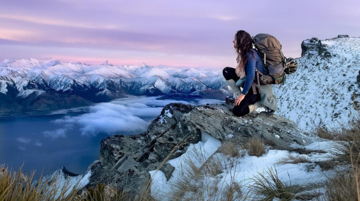 Na pierwszym planie na skale siedzi kobieta z plecakiem turystycznym wpatrująca się w krajobraz górski.