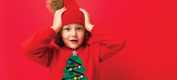 Na czerwonym tle wesoły chłopczyk w czerwonym swetrze z choinką i czerwonej czapce z pomponem trzyma się za głowę