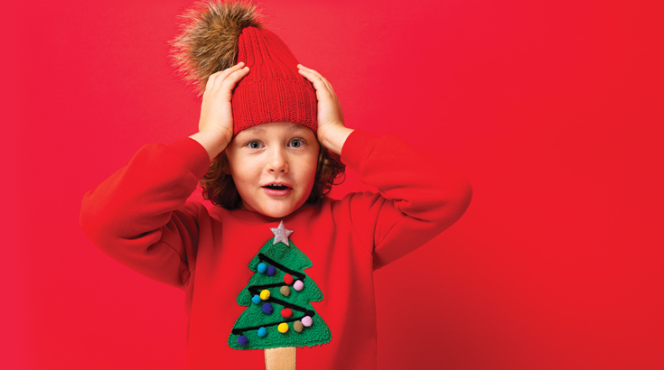 Na czerwonym tle wesoły chłopczyk w czerwonym swetrze z choinką i czerwonej czapce z pomponem trzyma się za głowę