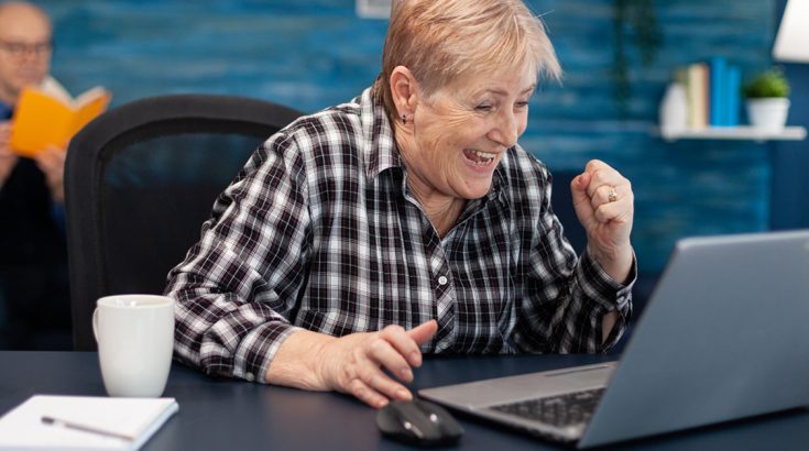 Seniorka z krótkimi blond włosami pochyla się nad ekranem laptopa śmiejąc się i ściskając pięść w geście zwycięstwa.