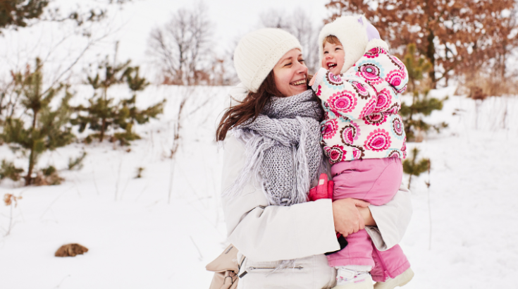 Roześmiana mama trzyma córkę na rękach na tle zimowego krajobrazu.