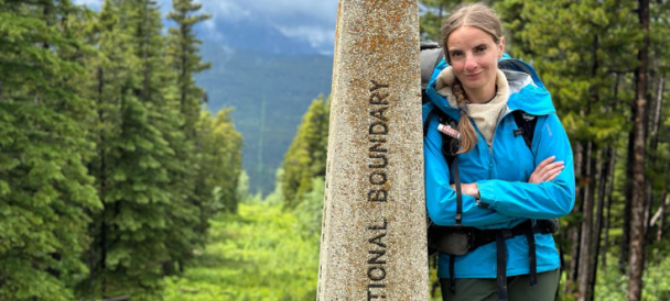 Po prawej stronie fotografii kobieta oparta o kamienny posąg wyznaczający szlak Pacific Northwest Trail. W tle widok na góry i las.