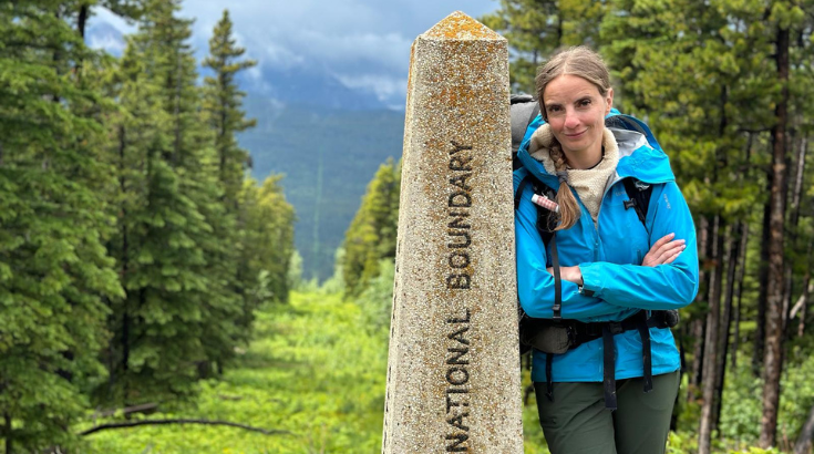 Po prawej stronie fotografii kobieta oparta o kamienny posąg wyznaczający szlak Pacific Northwest Trail. W tle widok na góry i las.