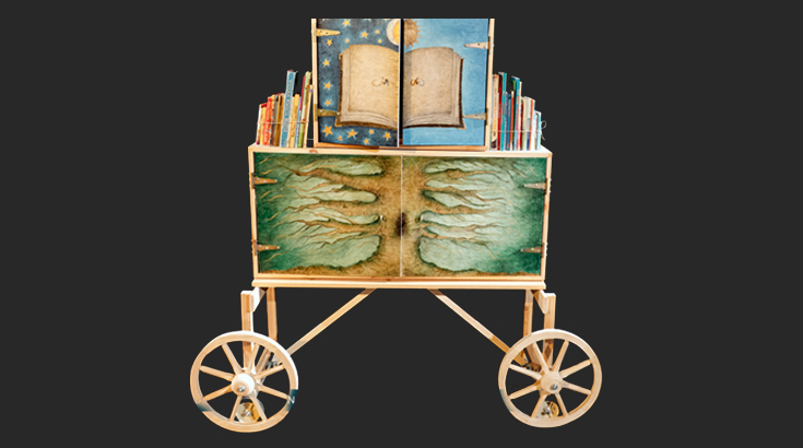 Na czarnym tle drewniany wóz przypominający objazdowe teatrzyki lalkowe