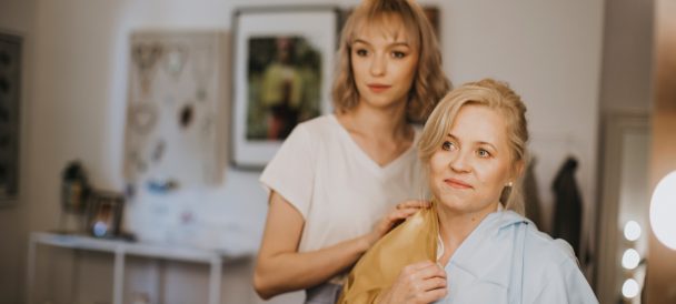 Dwie kobiety patrzą w stronę obiektywu jak w lustro. Jedna siedzi a druga przykłada jej złoty szal do twarzy i ubrania dopasowując.