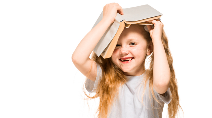 Na białym tle uśmiechnięta dziewczynka z książką na głowie.
