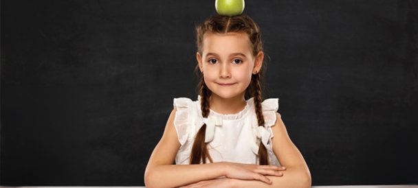 Na czarnym tle siedzi dziewczynka w białej bluzce z dwoma warkoczami z założonymi rękoma opartymi o biały blat. Na głowie ma zielone jabłko.