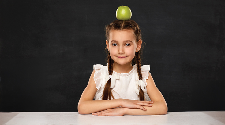 Na czarnym tle siedzi dziewczynka w białej bluzce z dwoma warkoczami z założonymi rękoma opartymi o biały blat. Na głowie ma zielone jabłko.